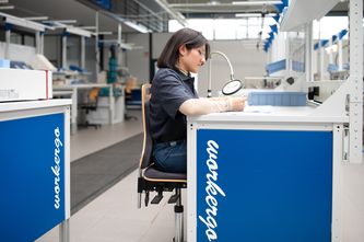 Eine Mitarbeiterin sitzt an einem ergonomischen Arbeitsplatz und prüft mit Hilfe einer Lupe ein Arbeitsstück
